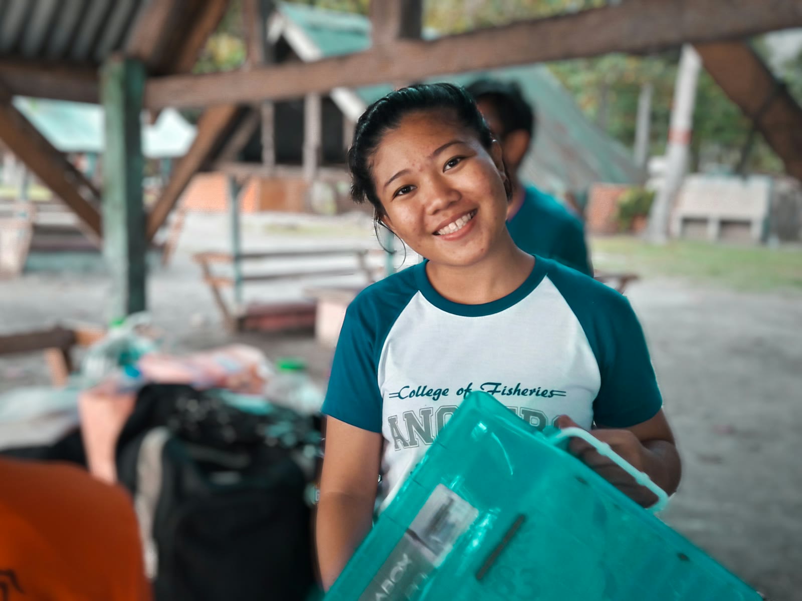 A smiling young volunteer carrying a crate of supplies at a community relief station under a pavilion roof