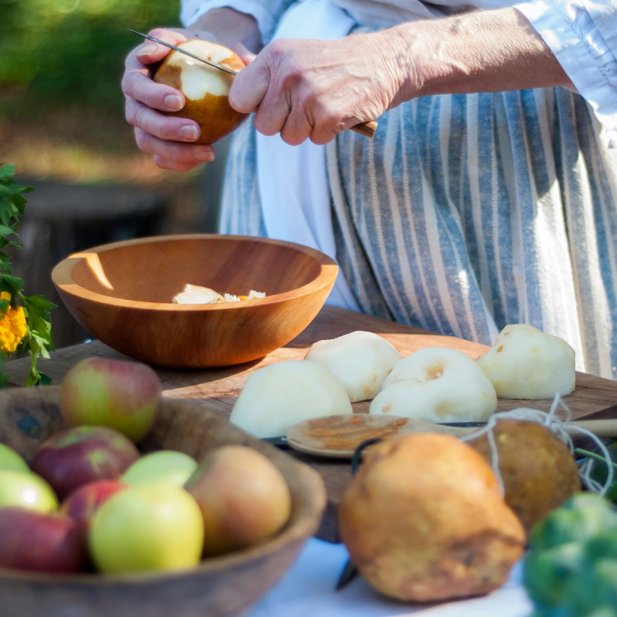 Hands peeling fruit at an outdoor table laid with apples, pears and fresh greens